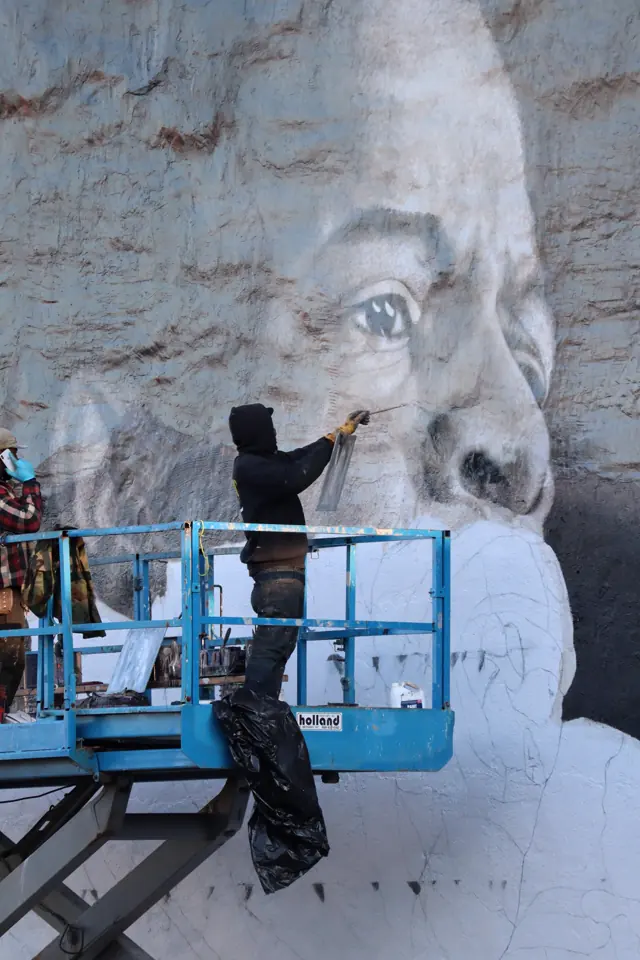 A painter on a lift at John Lewis’s eye, the rest of the face still in grid outline on the white wall